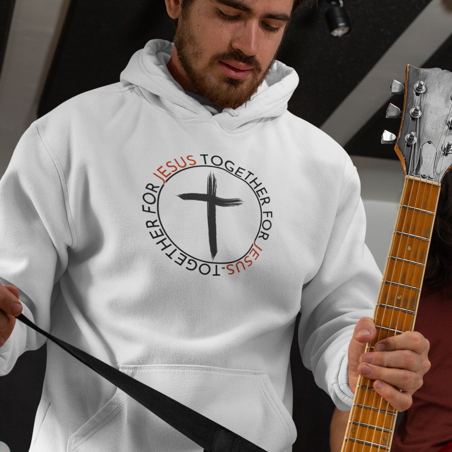 Man wearing a hoodie with a cross design and text, holding a guitar.