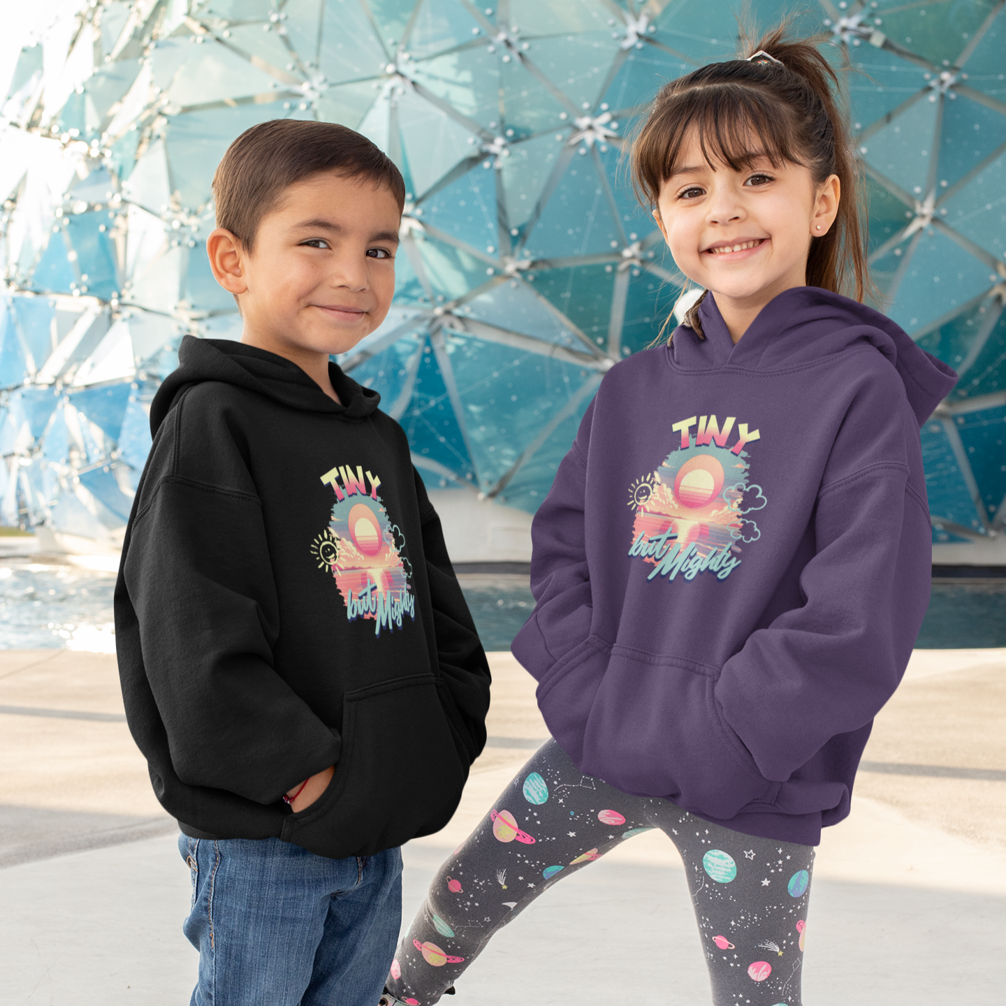 Two children wearing hoodies and colorful leggings standing in front of a textured wall.