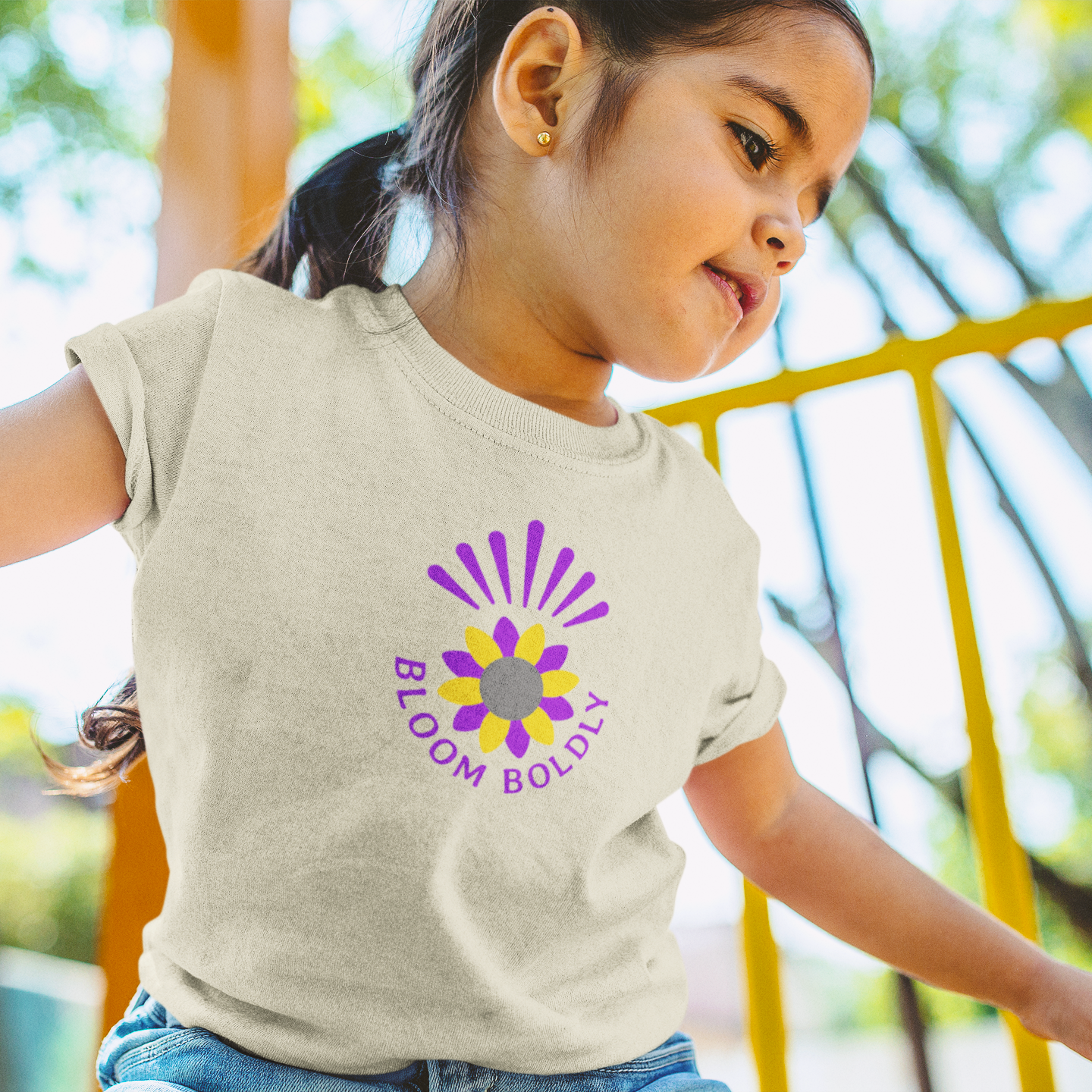 Child wearing a beige t-shirt with a purple and yellow 'Bloom Boldly' logo outdoors.