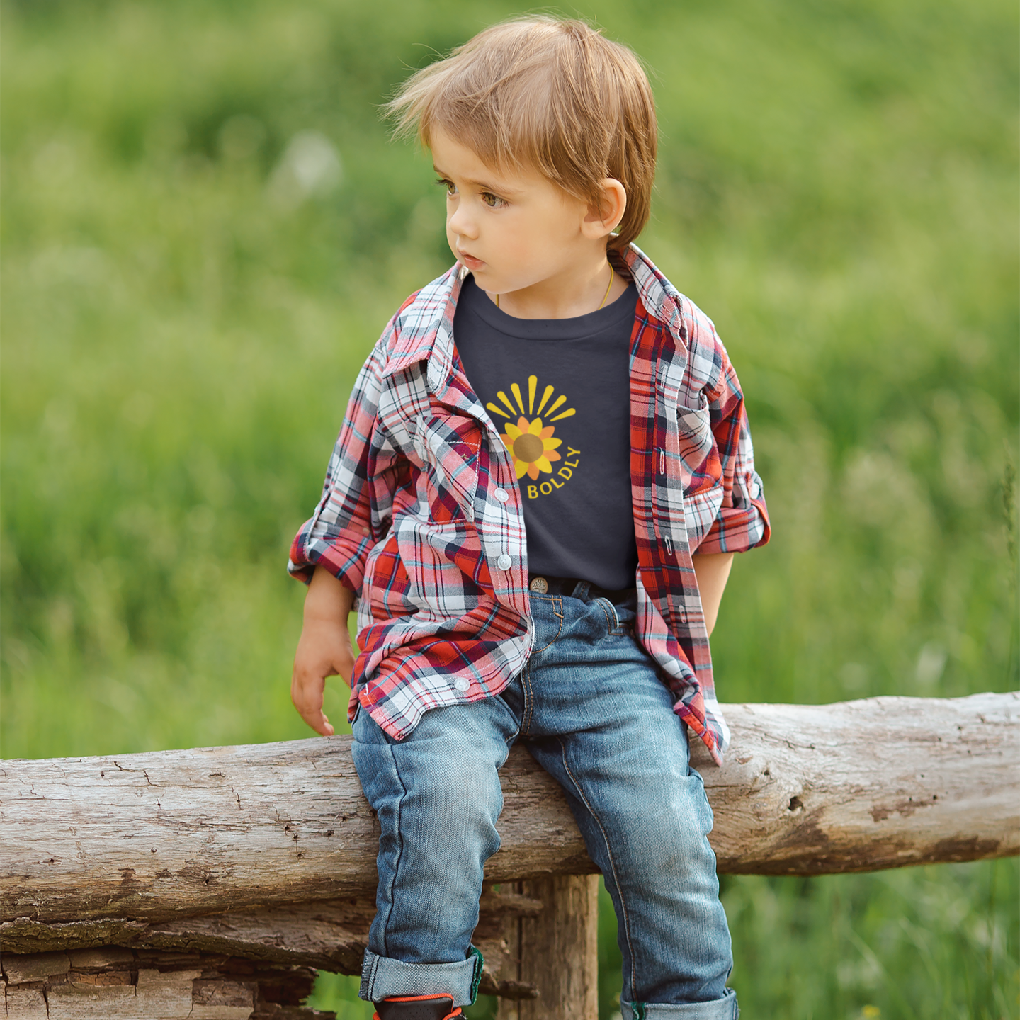 Child sitting on a log in a grassy field wearing a plaid shirt and jeans.