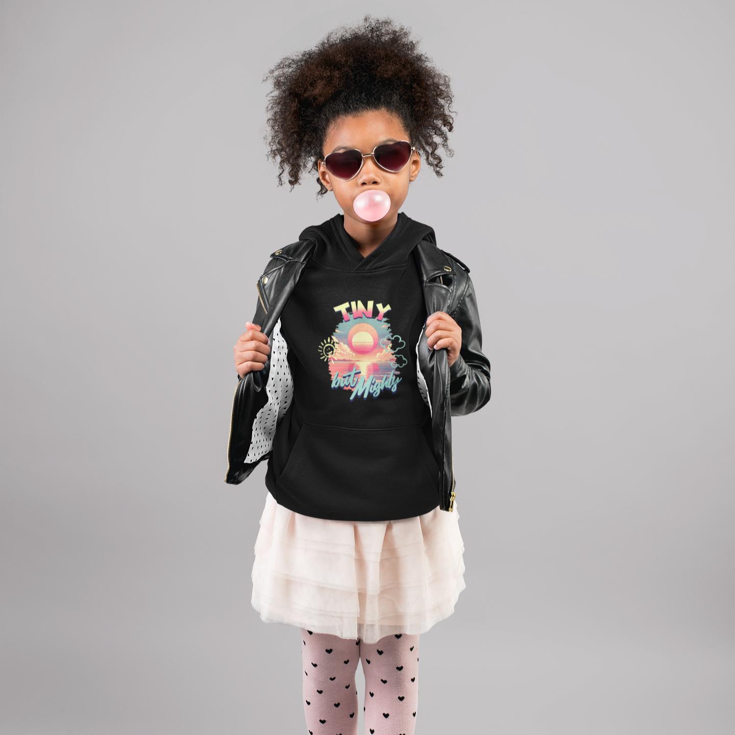Child wearing a black hoodie with a graphic design, sunglasses, and a leather jacket on a gray background